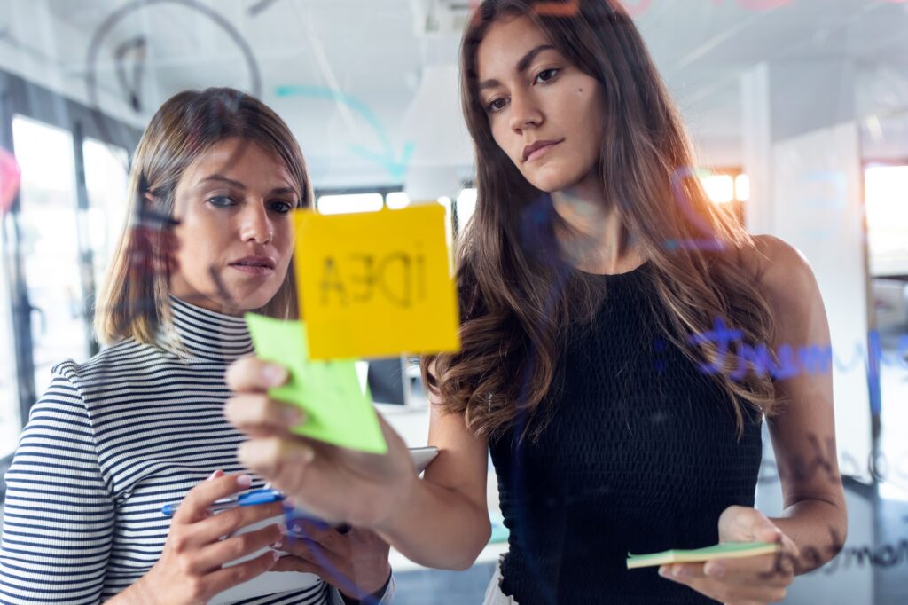 Two business young women working together on wall glass with post it stickers